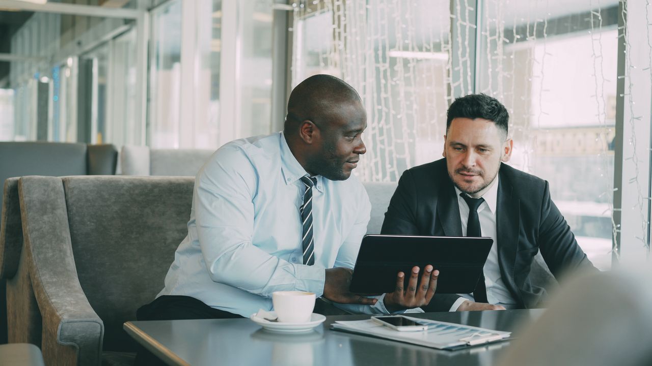 Two men in business attire collaborating over a tablet in a modern office setting.