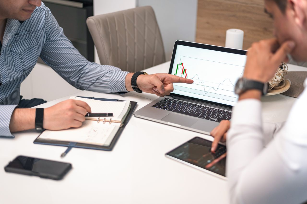 Two businessmen analyzing financial charts and data on a laptop and tablet.