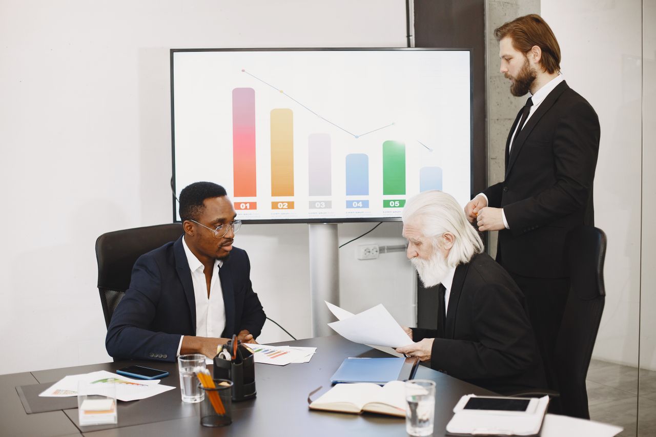 our-journey-03 Three businessmen discussing financial data in a modern office setting with charts.
